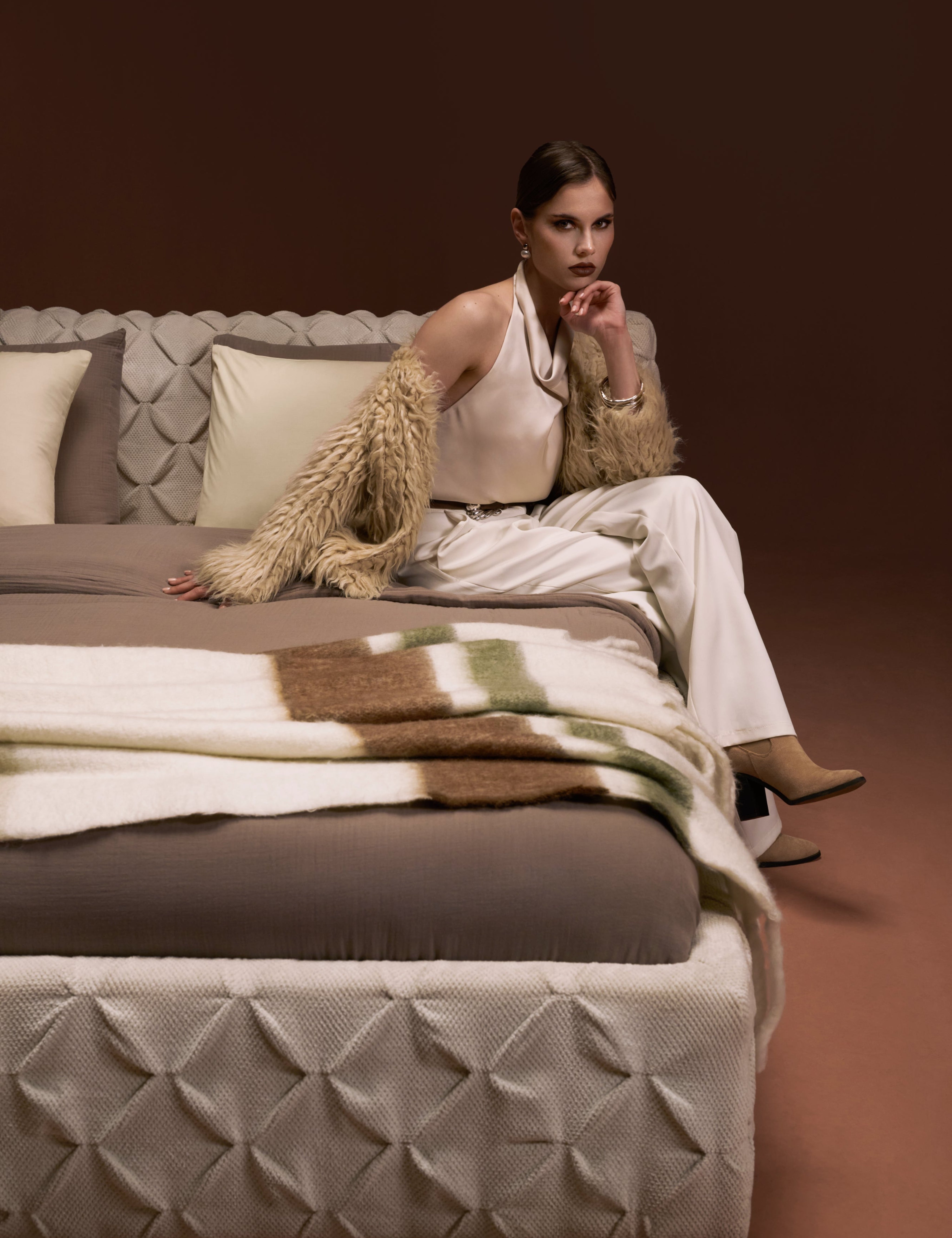 Model seated on Ro-Blocks bed with pinch pleated headboard and base, upholstered in cream fabric, shown in an elevation view.