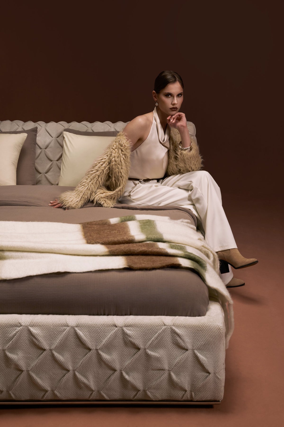 Model seated on Ro-Blocks bed with pinch pleated headboard and base, upholstered in cream fabric, shown in an elevation view.