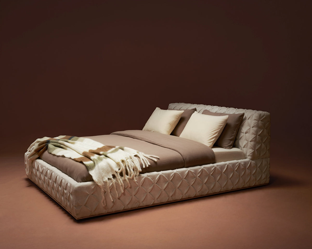 Wide side shot of the bed in cream fabric with pinch-pleated headboard and base.
