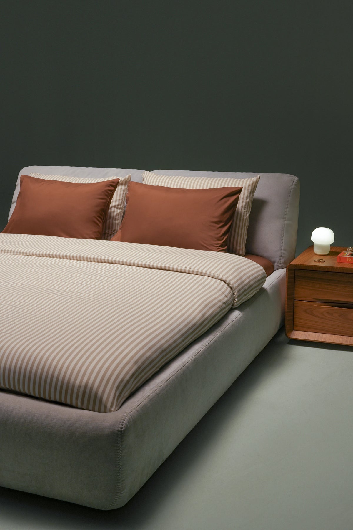 Snug upholstered bed with raised adjustable backrests, a recessed mattress and a wooden two-drawer nightstand with a small lamp.