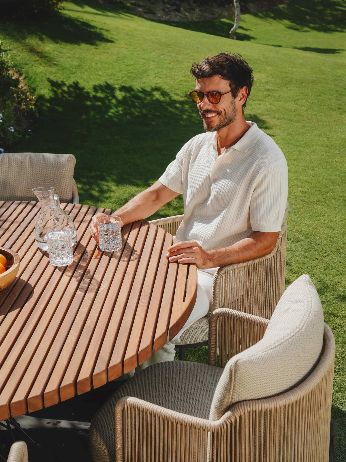 Man seated in a beige rope Cord dining chair at the round Spiral teak dining table on a sunny garden lawn.