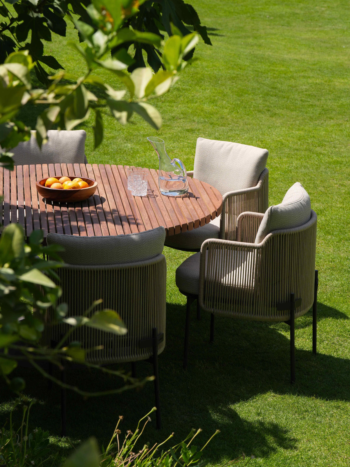 Spiral teak dining table with Cord dining chairs featuring beige handwoven rope frames and cushioned seats, arranged on a garden lawn.